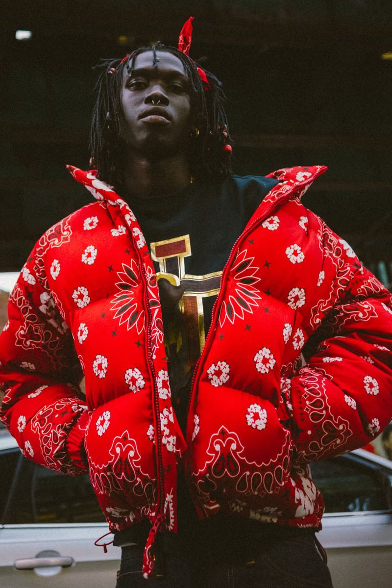 Peezy wears Denim Tears x Billionaire Boys Club collaboration pieces including chunky knit cardigan with red, black, and green striping on New York City street - detail view 1
