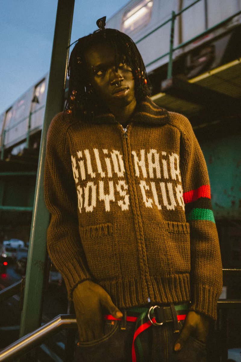 Peezy wears Denim Tears x Billionaire Boys Club collaboration pieces including chunky knit cardigan with red, black, and green striping on New York City street - detail view 2